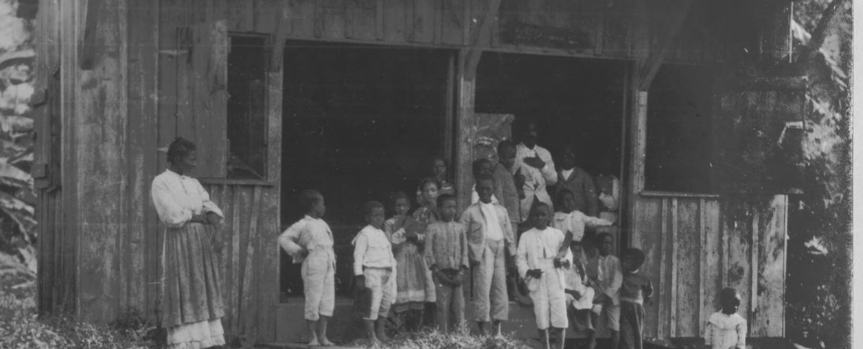 A group of young Jamaican children standing in front of a building, possibly a schoolhouse.